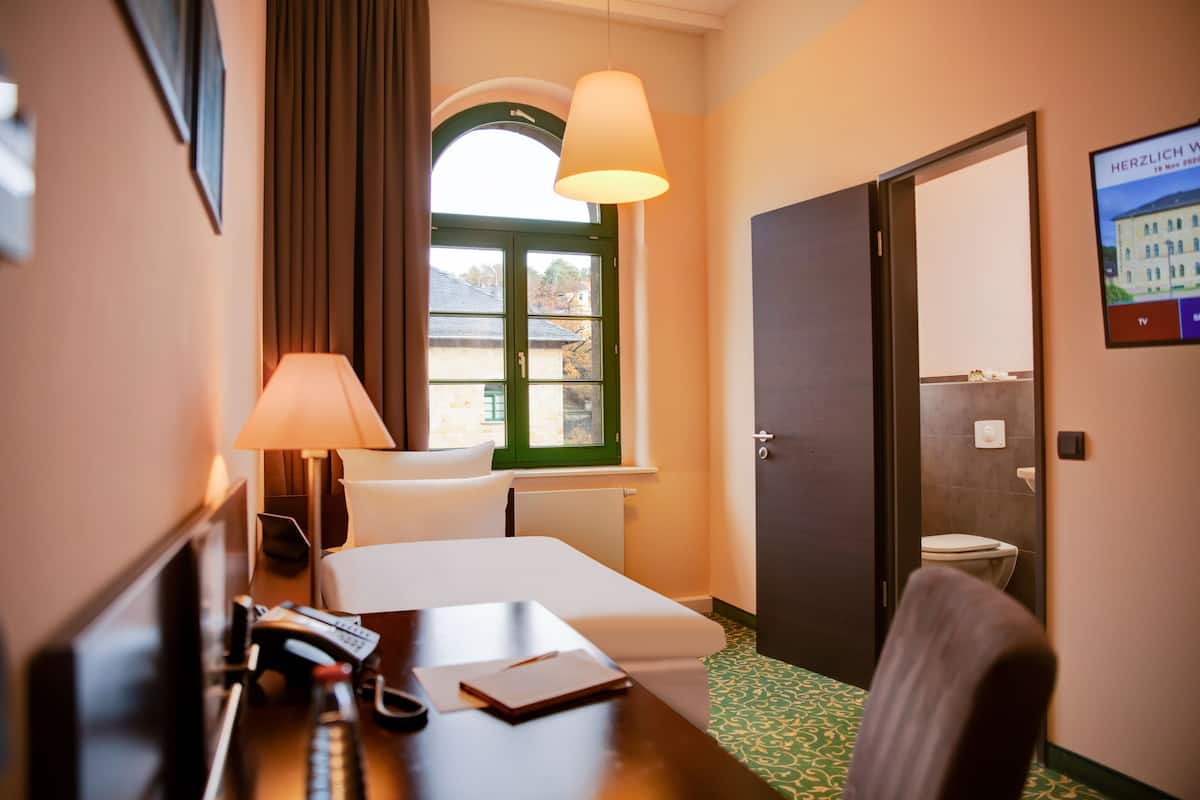 Habitación de hotel con cama, escritorio con lámpara, silla, ventana con cortinas y puerta que da a un baño.