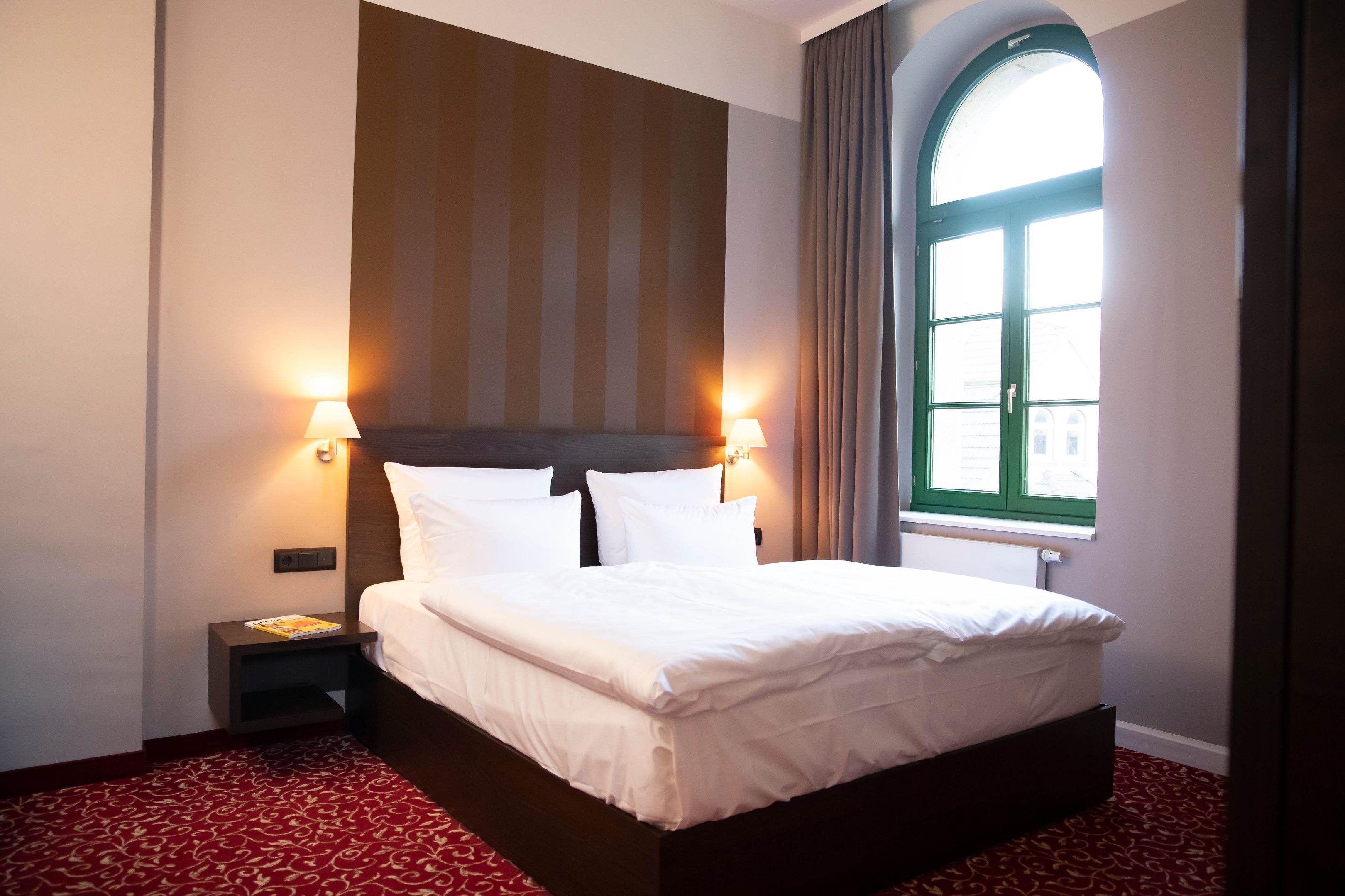Une chambre d’hôtel avec un grand lit, deux lampes de chevet, une table de nuit, une fenêtre avec des rideaux et un tapis rouge à motifs.