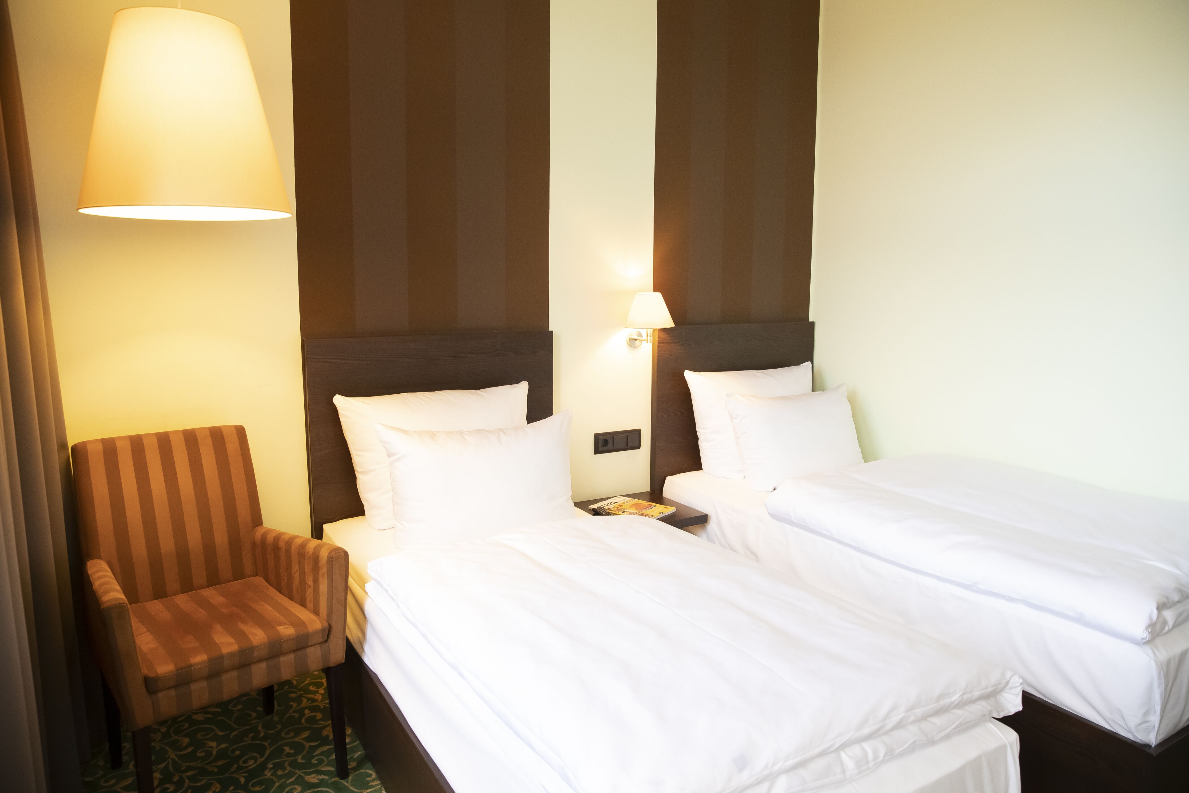 Camera d'albergo con due letti singoli, una sedia e una lampada.