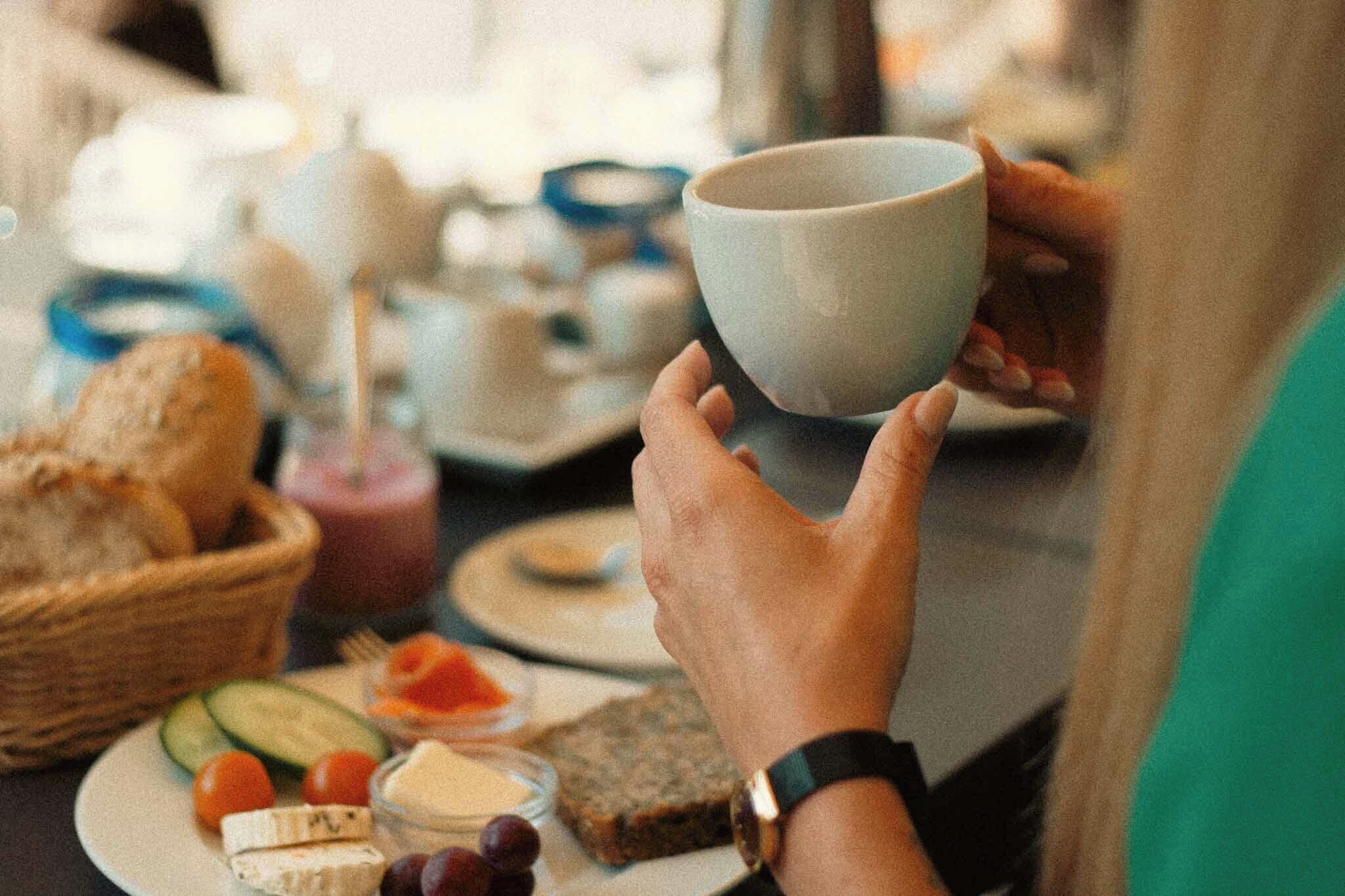 A person holding a cup, with a plate of food and a basket of bread in the foreground.