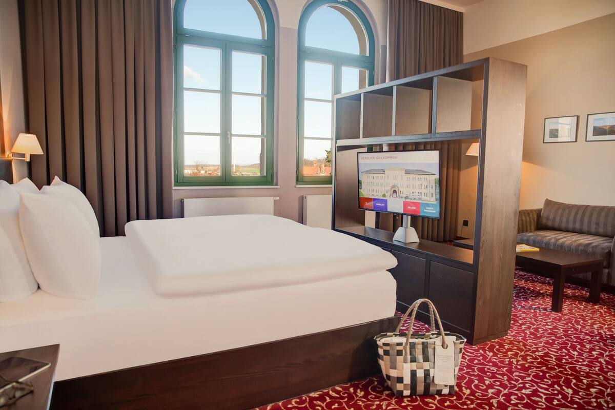 Habitación de hotel con una cama grande, un escritorio con computadora, una silla y vistas a la ciudad a través de amplias ventanas.