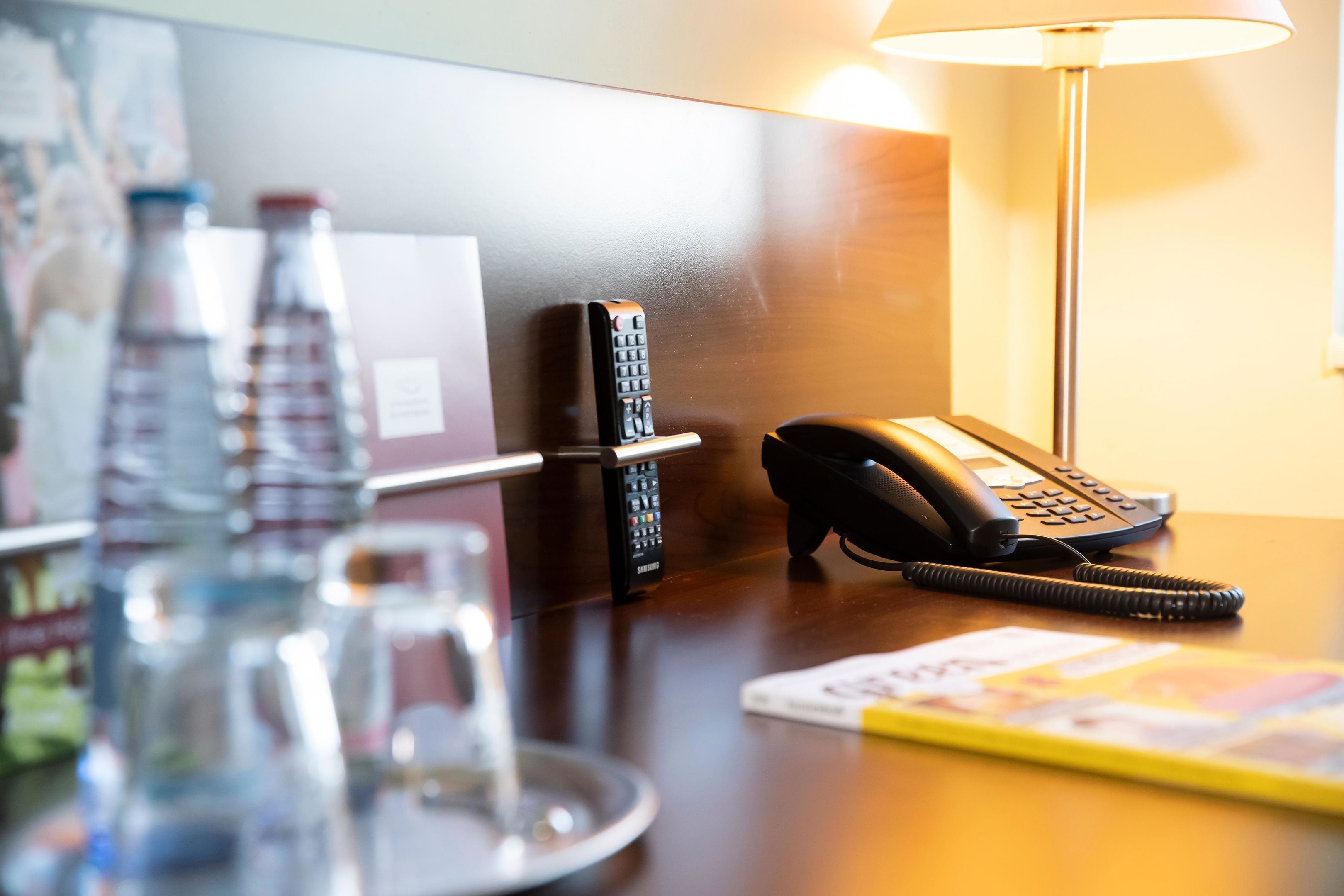Ein Schreibtisch im Hotelzimmer mit Telefon, Fernbedienung, Glas und Zeitschrift.