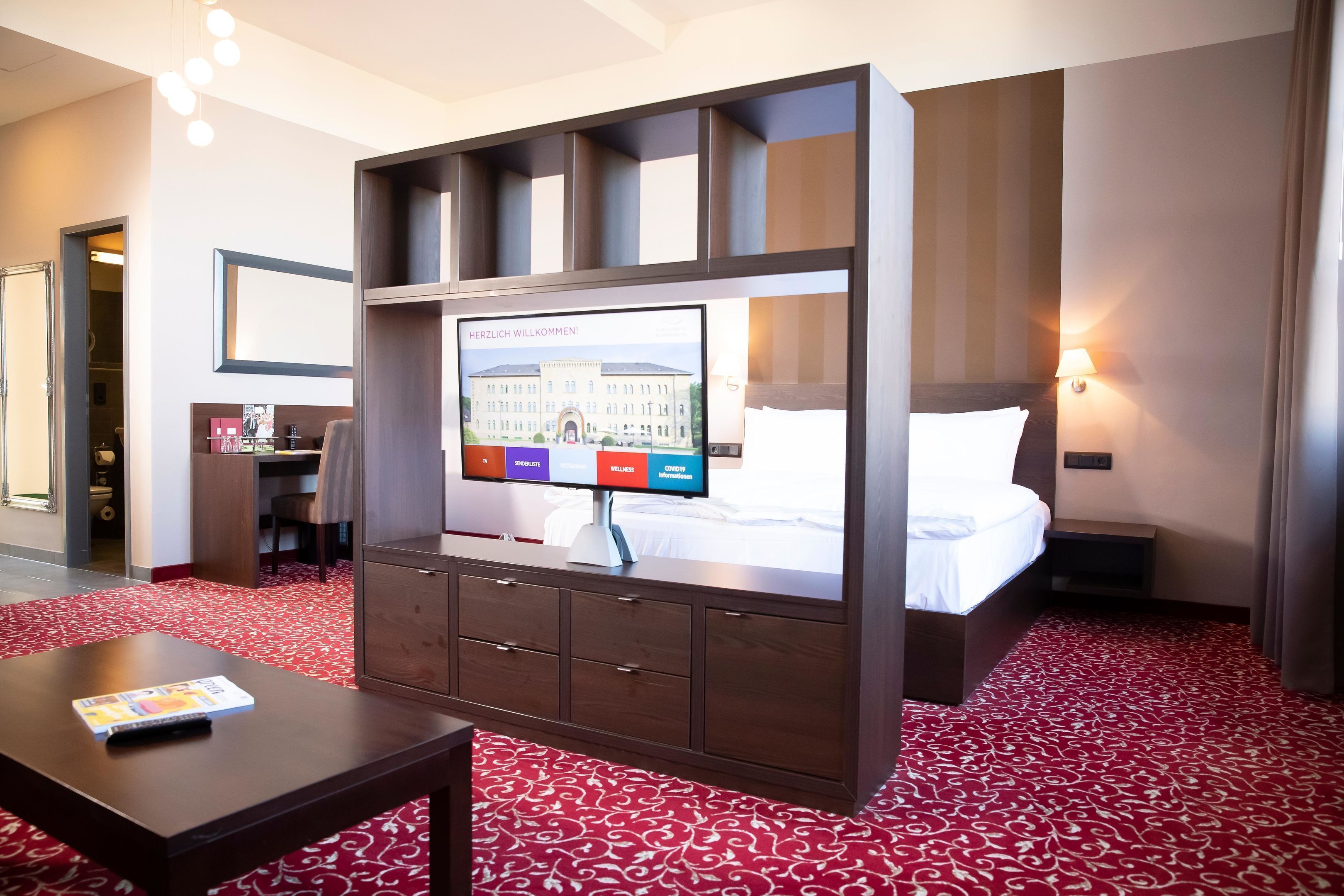 Hotelkamer met een bed, een bureau met een computer, een televisie bevestigd aan een houten kast en een tapijtel van rood dessin.
