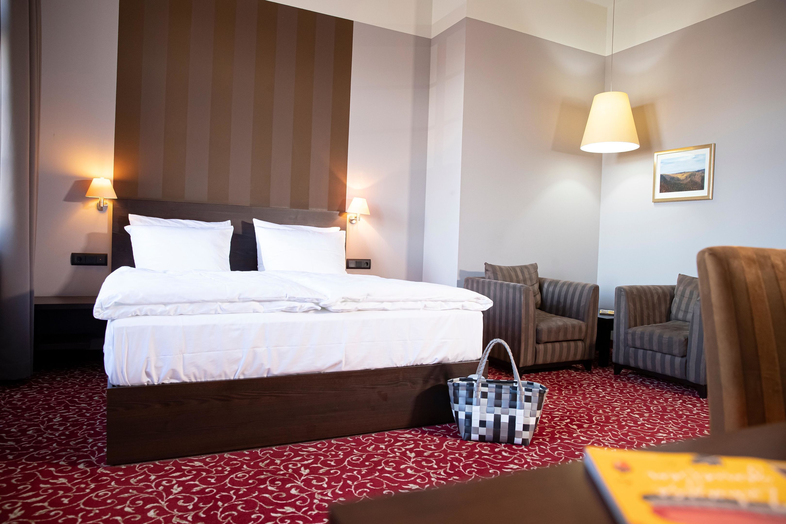 Habitación de hotel con una cama grande, dos sillones, una mesita pequeña y una alfombra roja estampada.
