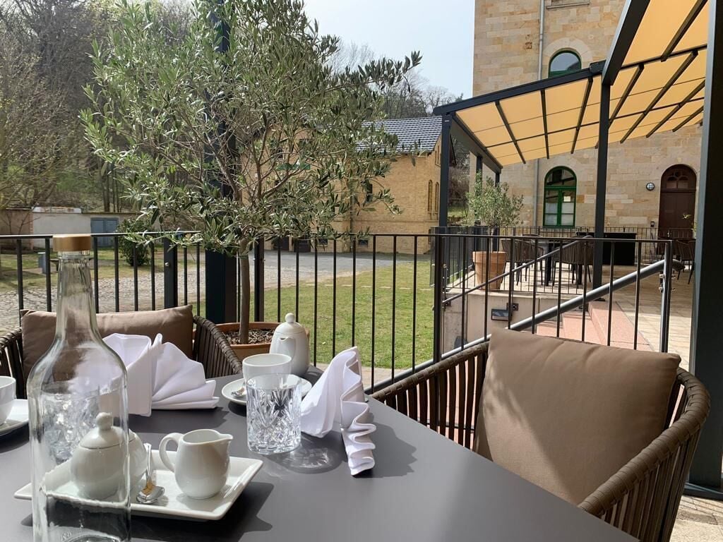 A table set for two with a bottle, teapot, cups, and a glass pitcher on a patio with a view of a building and trees.