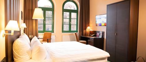 Habitación de hotel con cama, dos ventanas con cortinas, escritorio con lámpara, armario y televisión en la pared.