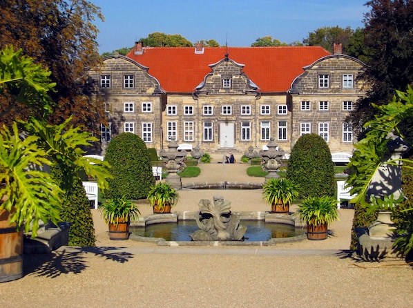 Un edificio grande e histórico con fachada simétrica, una entrada central y un jardín formal con una fuente.