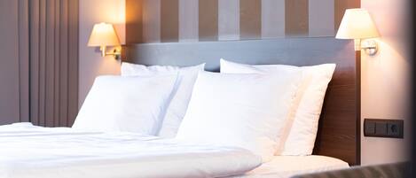 Een net opgemaakt bed met witte lakens en kussens, een houten hoofdbord en twee wandlampen aan weerszijden.