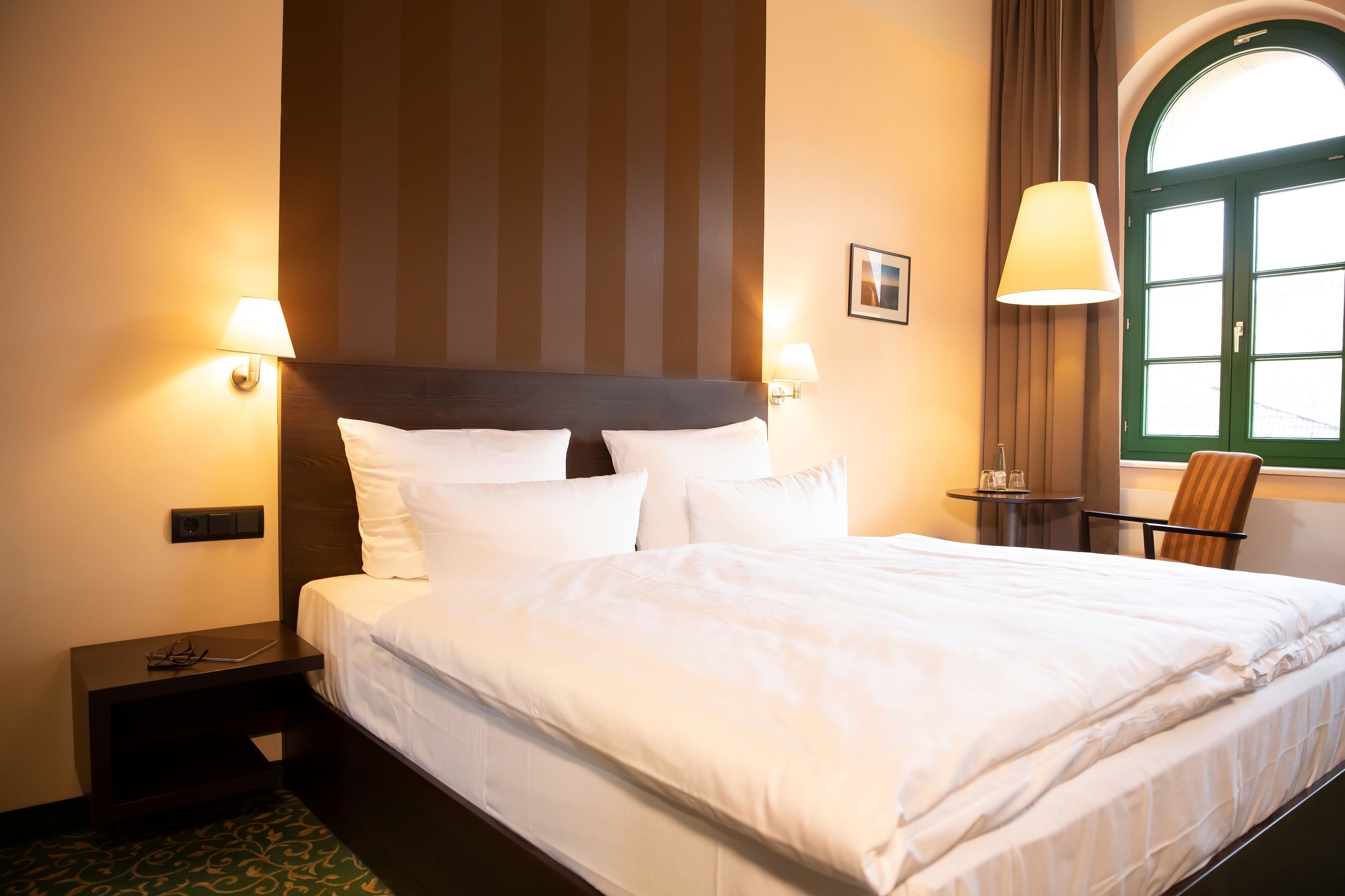 Een hotelkamer met een groot bed, twee bedlampen, een kleine tafel met een stoel en een raam met gordijnen.