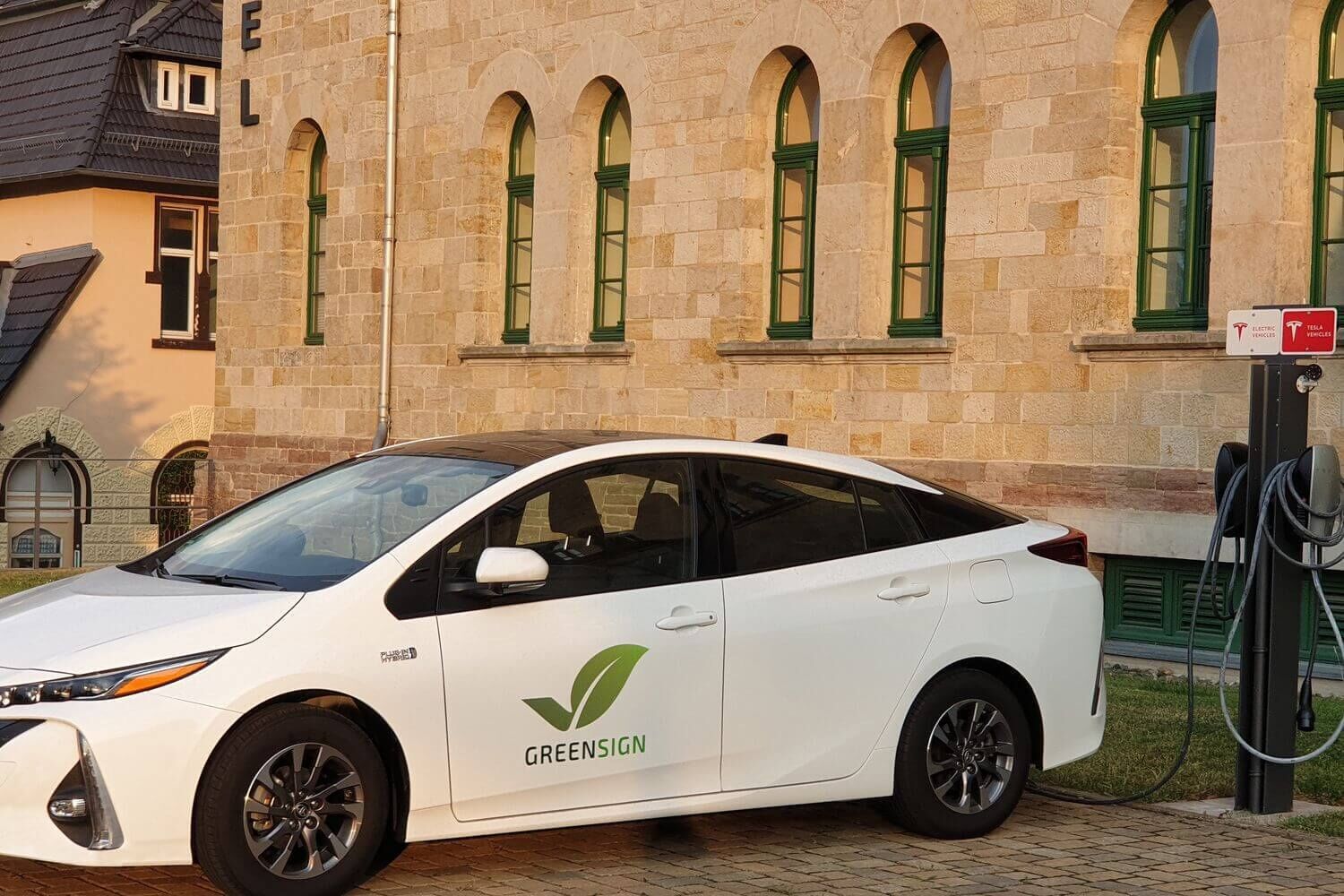 Ein weißes Elektroauto mit dem Aufdruck "GREENSIGN" vor einem Steinhaus mit grünen Fensterrahmen.