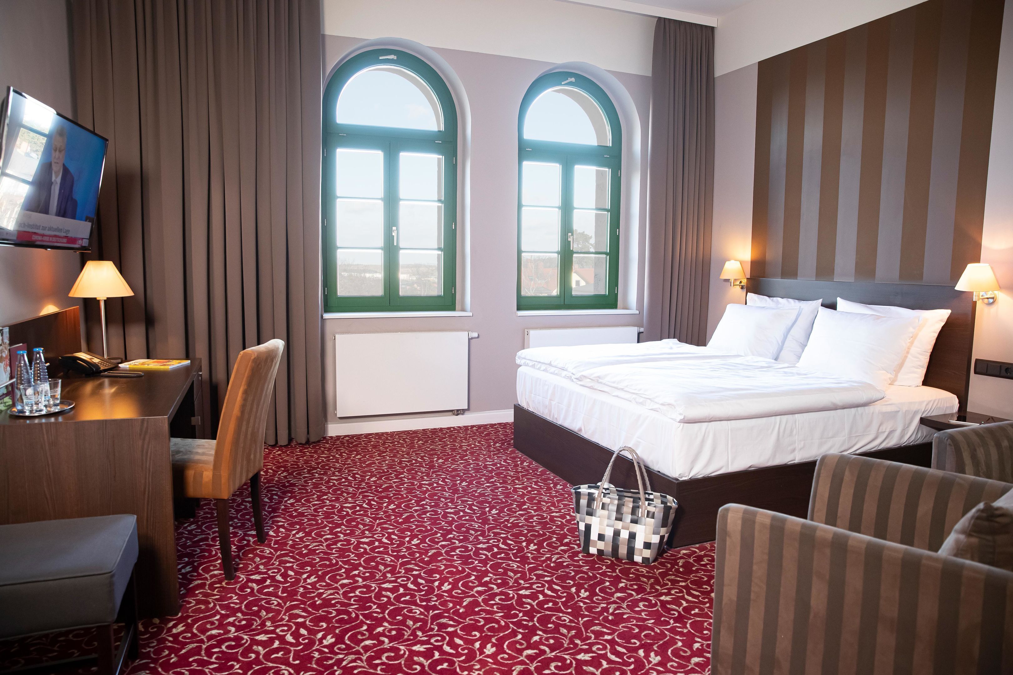 Ein Hotelzimmer mit einem Bett, zwei Fenstern, einem Schreibtisch, einem Stuhl und einem Fernseher.