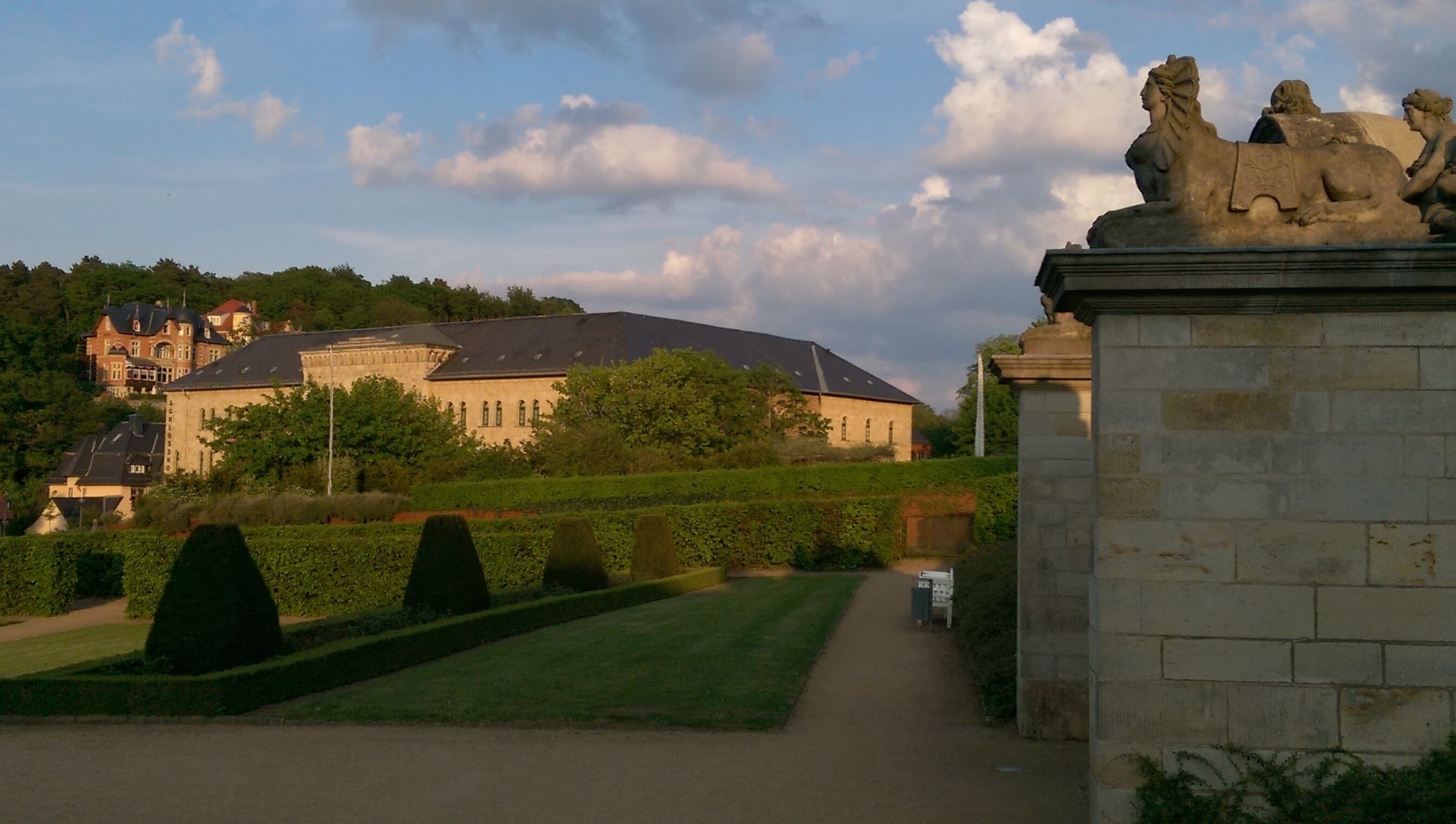 A pathway leading to a large building with a sloped roof, surrounded by well-manicured hedges and a statue.