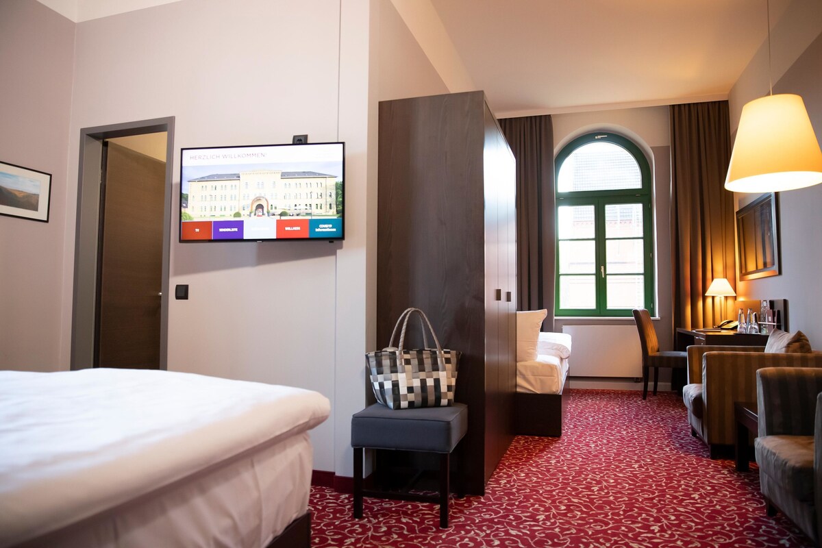 A hotel room with a bed, a desk, a chair, a TV mounted on the wall, and a window with curtains.