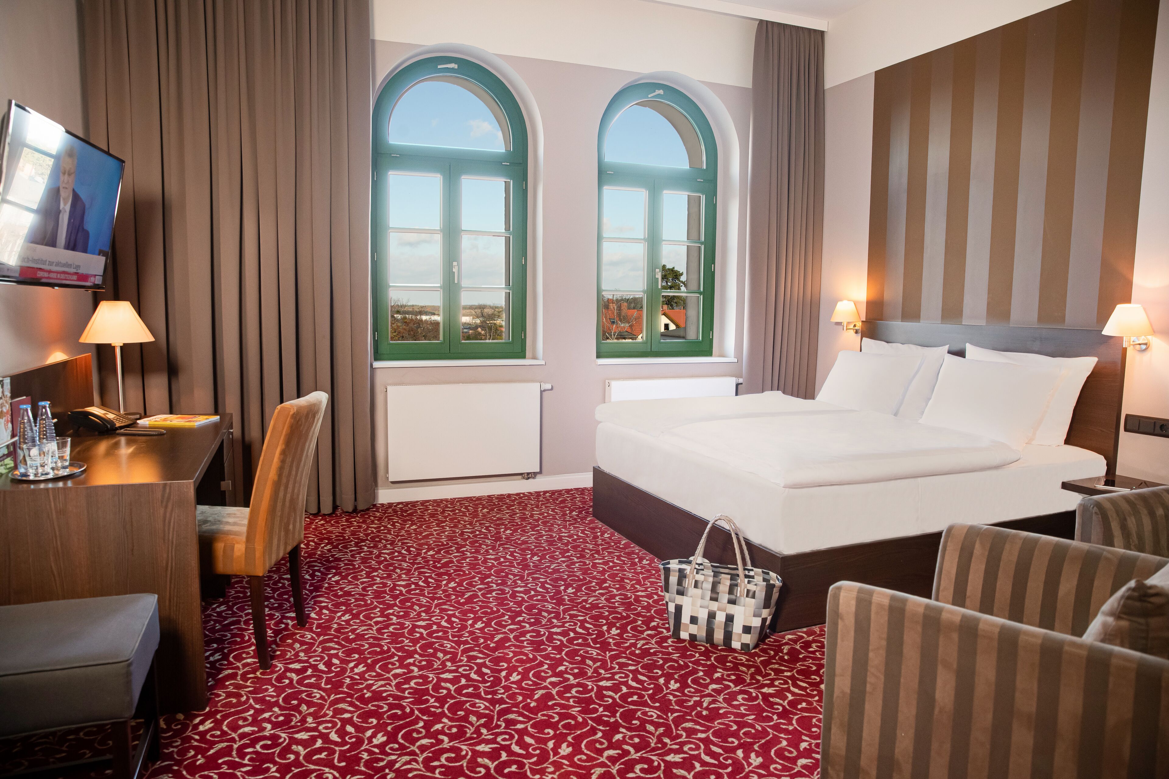 A hotel room with a large bed, two windows with curtains, a desk with a chair, and a TV on the wall.