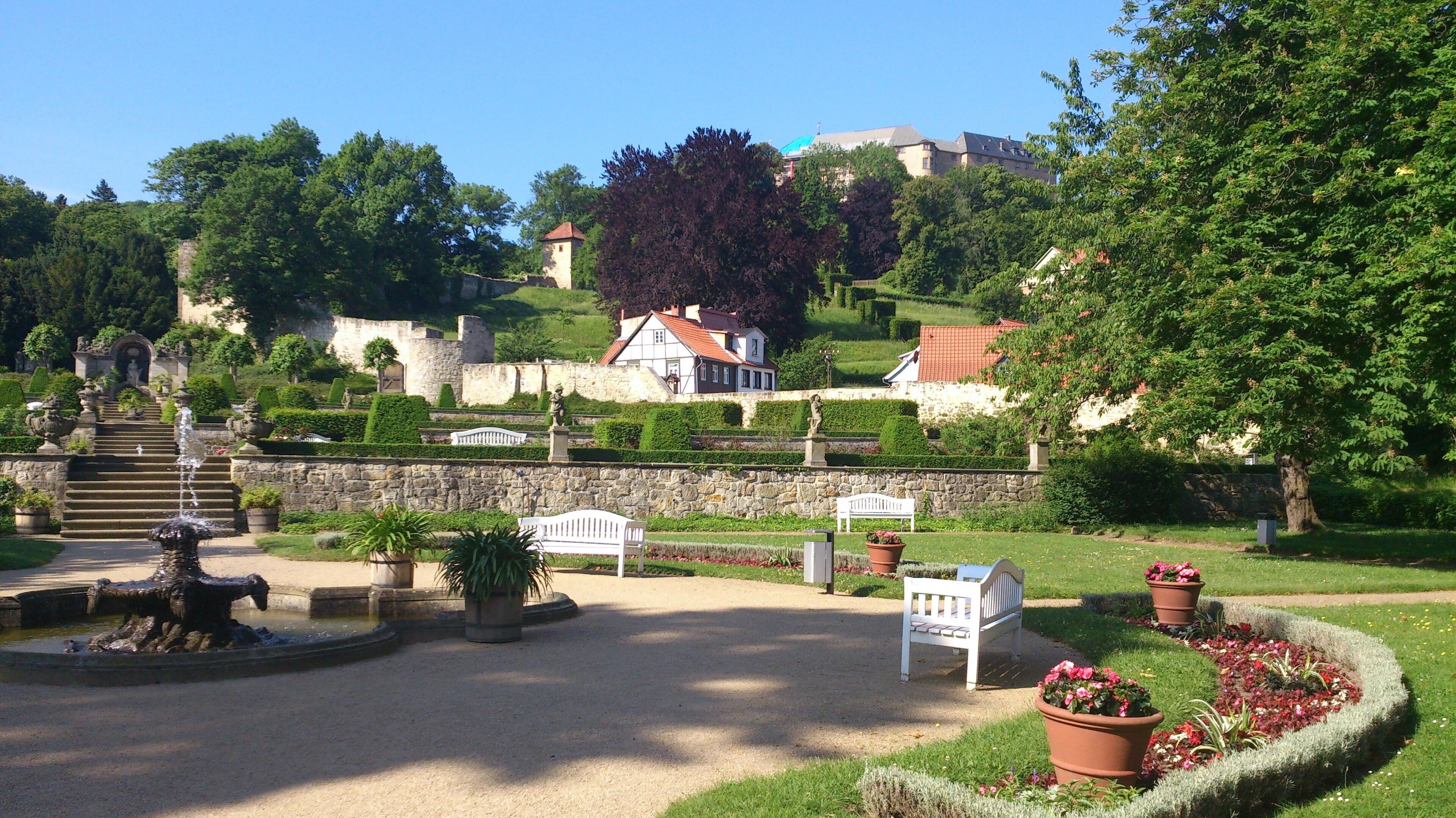 A garden with a fountain, benches, and a statue. There are stone walls and a building in the background.