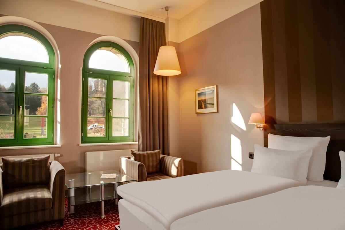 Habitación de hotel con una cama grande, dos sillones, una mesita y vistas a la vegetación a través de ventanas con marcos verdes.