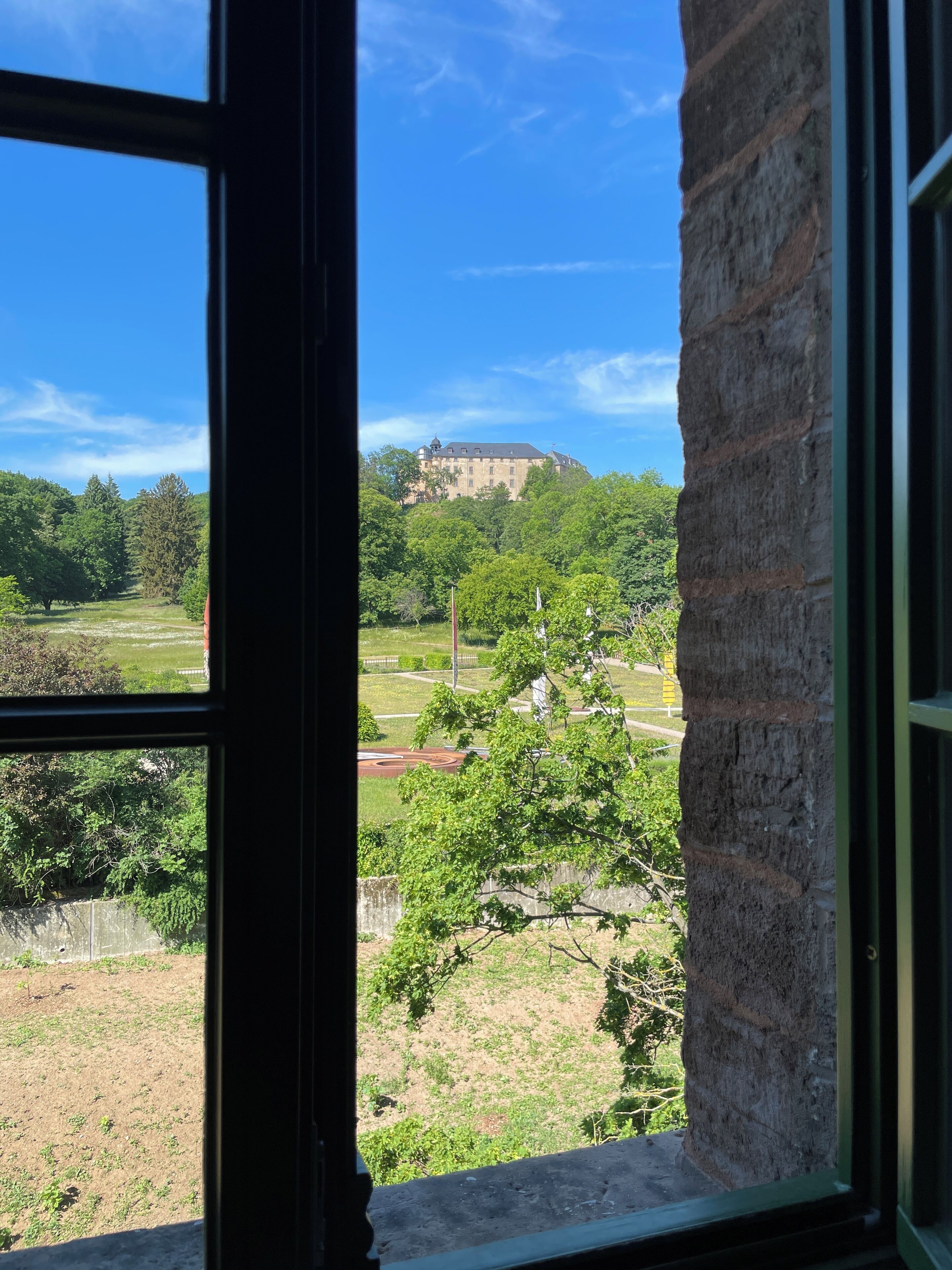 Vista attraverso una finestra su un edificio con un muro in pietra, una zona verdeggiante e una struttura distante simile a un castello.