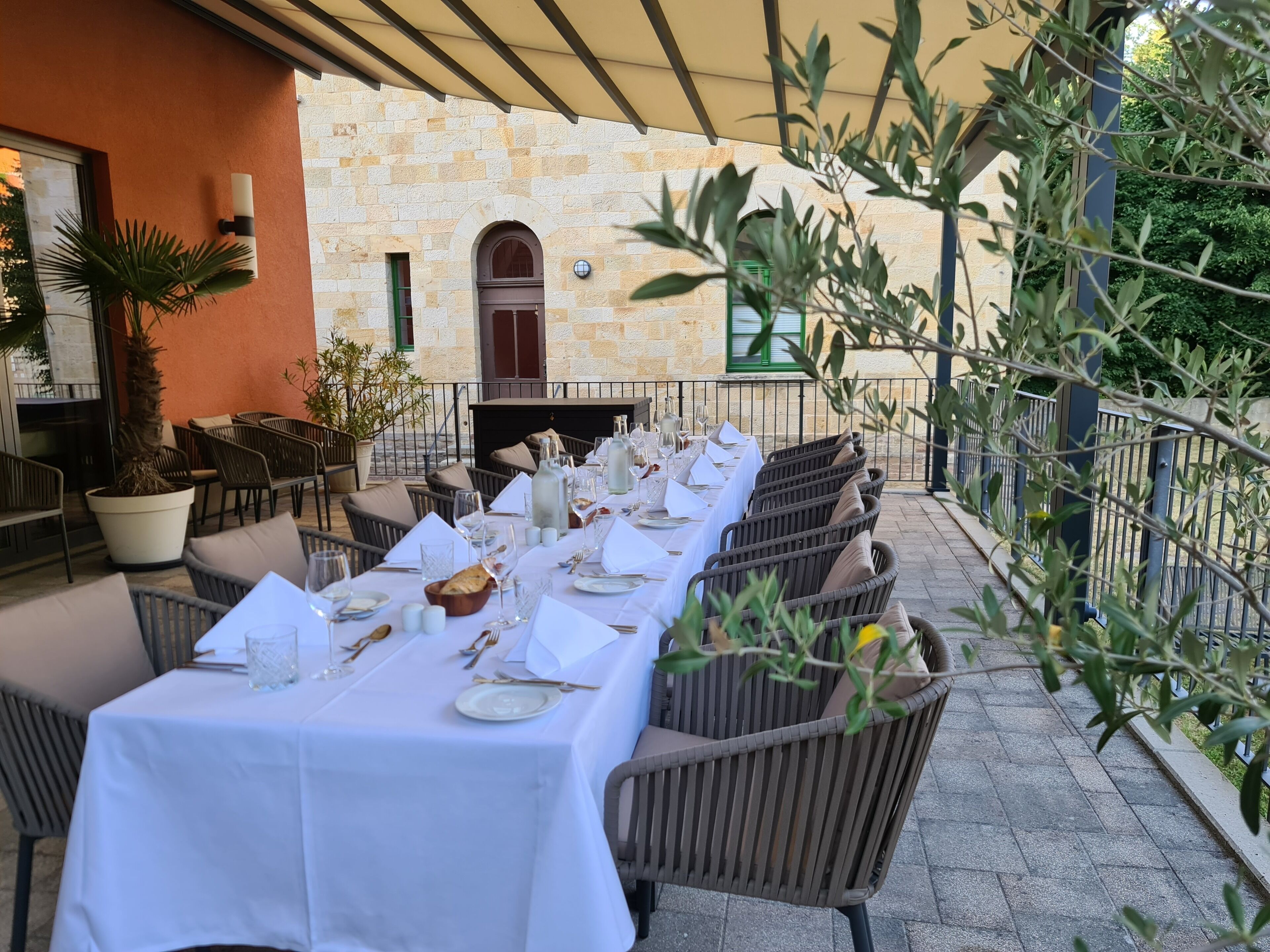 A long dining table set with white tablecloths, plates, and cutlery, surrounded by wicker chairs.