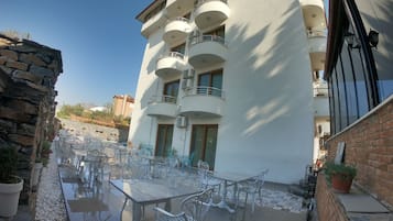 Un edificio con varios balcones, zona de estar al aire libre con mesas y sillas, y un muro de piedra en un lateral.