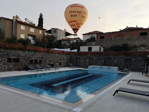 A swimming pool with lounge chairs, a stone wall, and a hot air balloon in the background. - Bellamaritimo Hotel (Denizli)