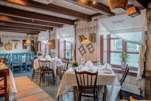 Un restaurante con mesas y sillas de madera, manteles blancos, faroles colgantes y vista a la calle.