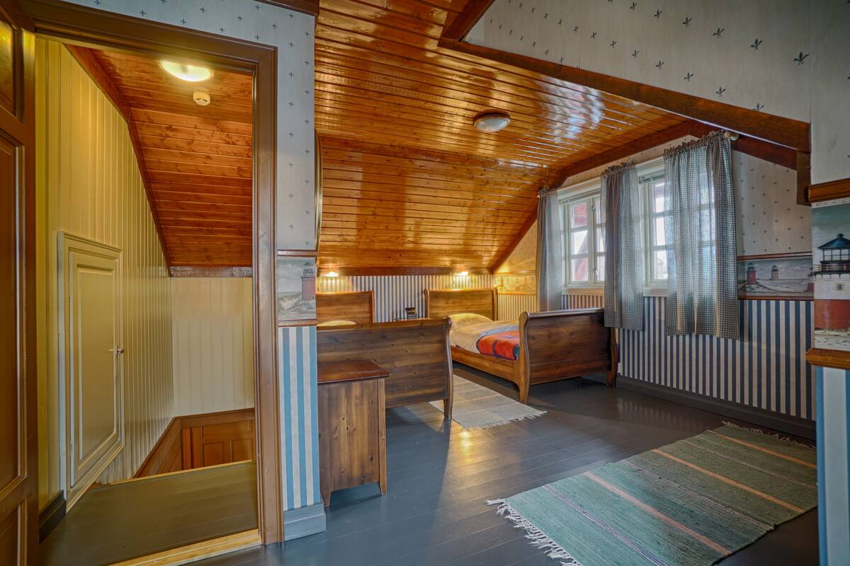 A cozy attic bedroom with a sloped ceiling, wooden beams, a bed, a nightstand, and a window with curtains.