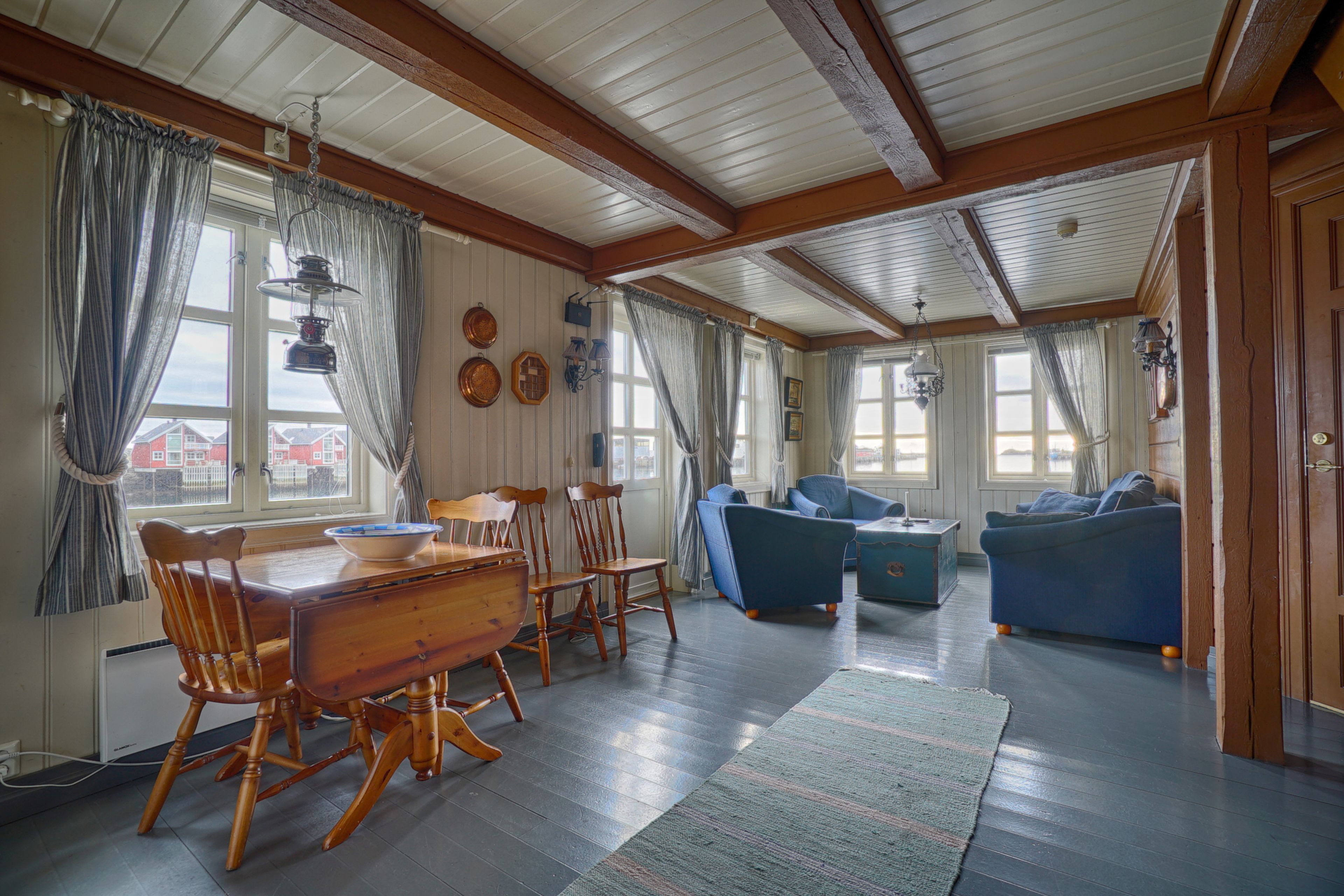 A spacious living room with a wooden dining table and chairs, a blue sofa, and a large window with curtains.