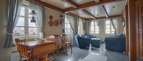 A spacious living room with a wooden dining table and chairs, a blue sofa, and a large window with curtains.