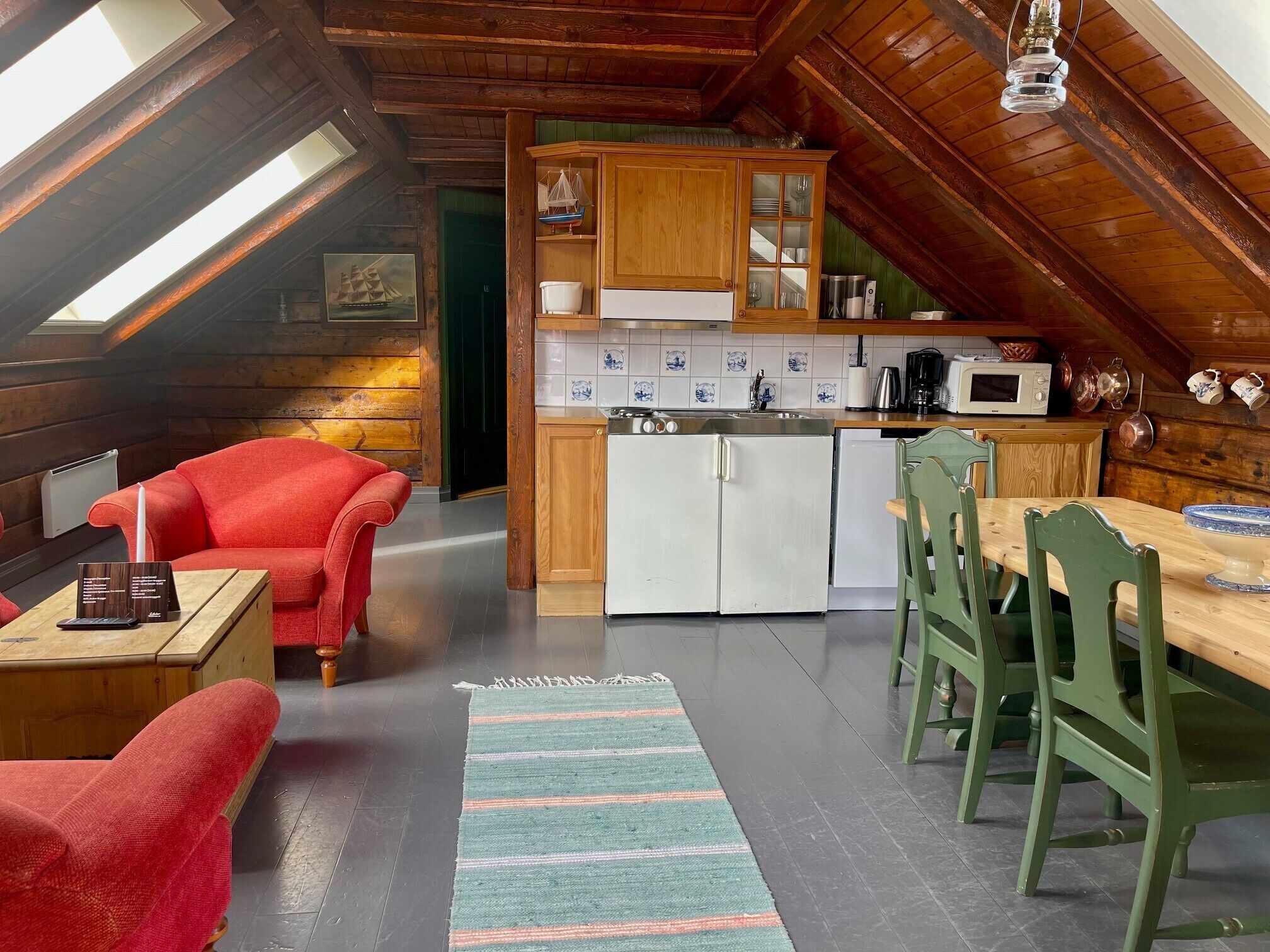 A cozy attic room with a kitchenette, red upholstered chairs, a wooden dining table, and a striped rug.