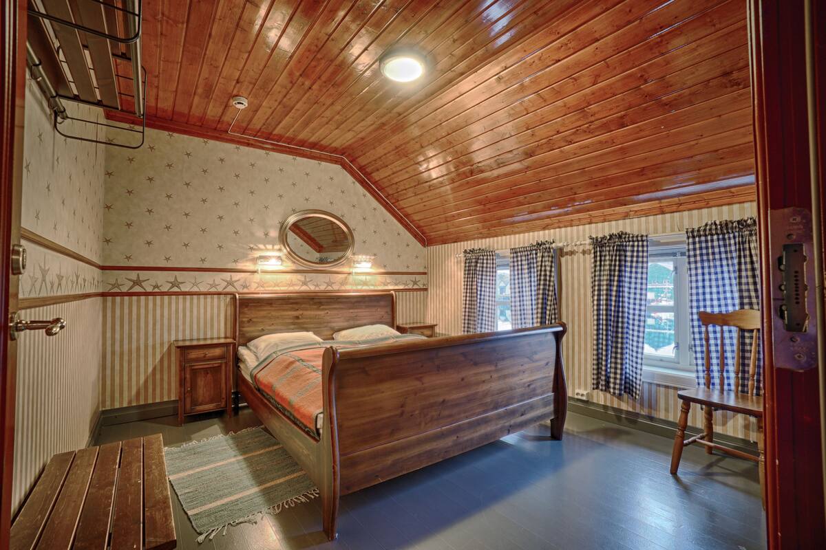 A bedroom with a wooden ceiling, a bed, a nightstand, a mirror, and a window with checkered curtains.
