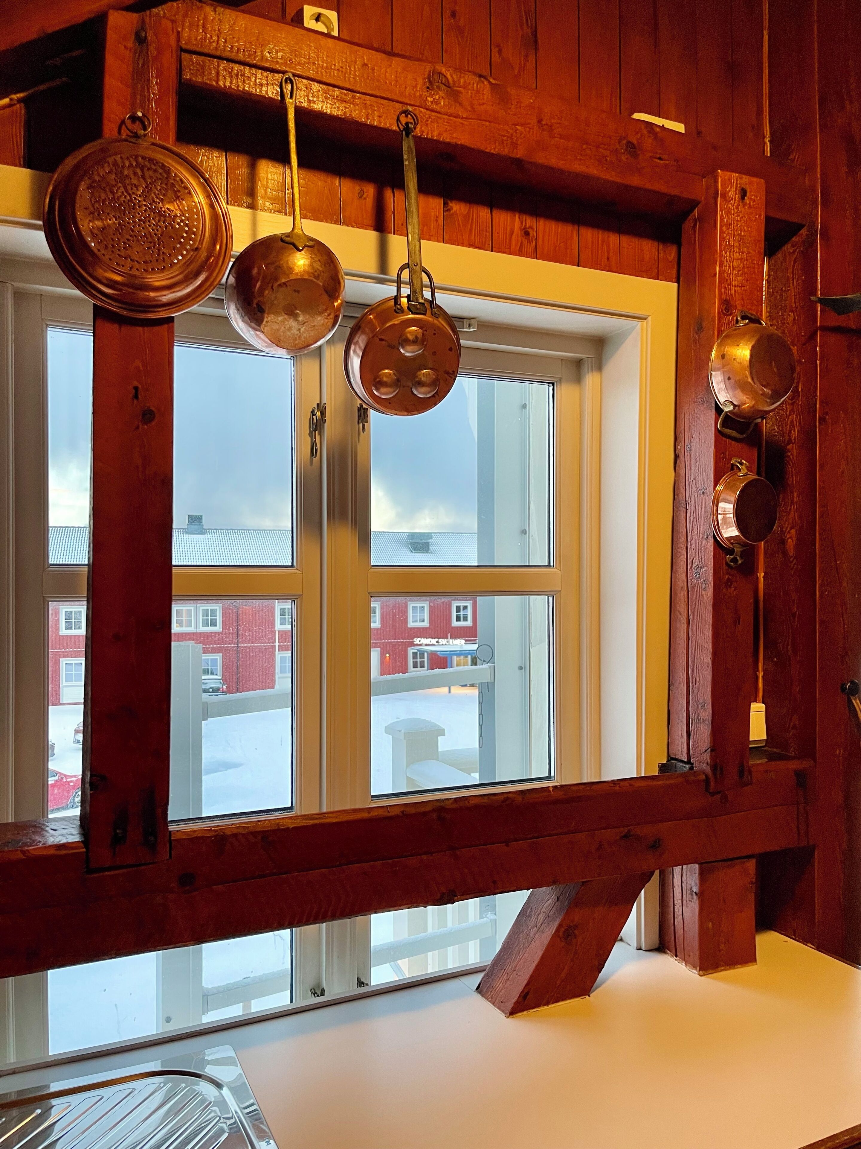 A kitchen with copper pots hanging from the ceiling, a window with a view of buildings, and wooden walls.