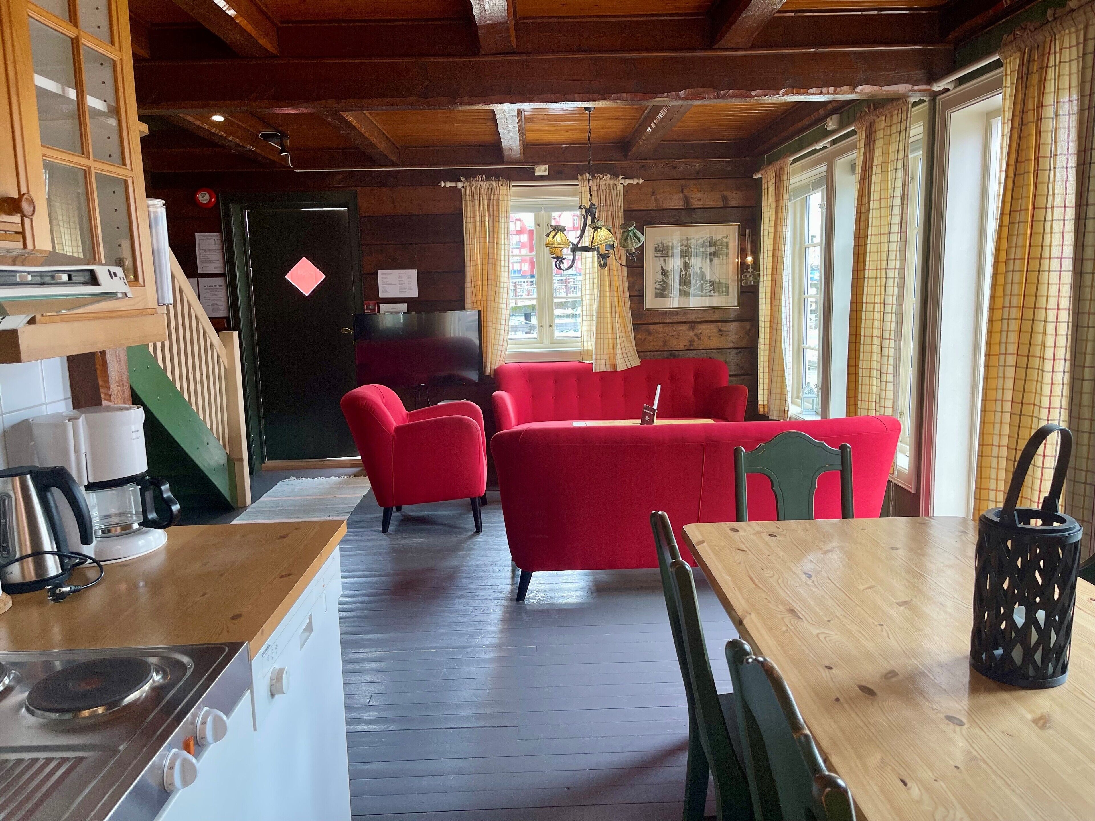 Ein gemütliches Zimmer mit Holzbalkendecke, einer roten Couch, einem Holztisch und einer Küchenzeile mit Herd und Kaffeemaschine.