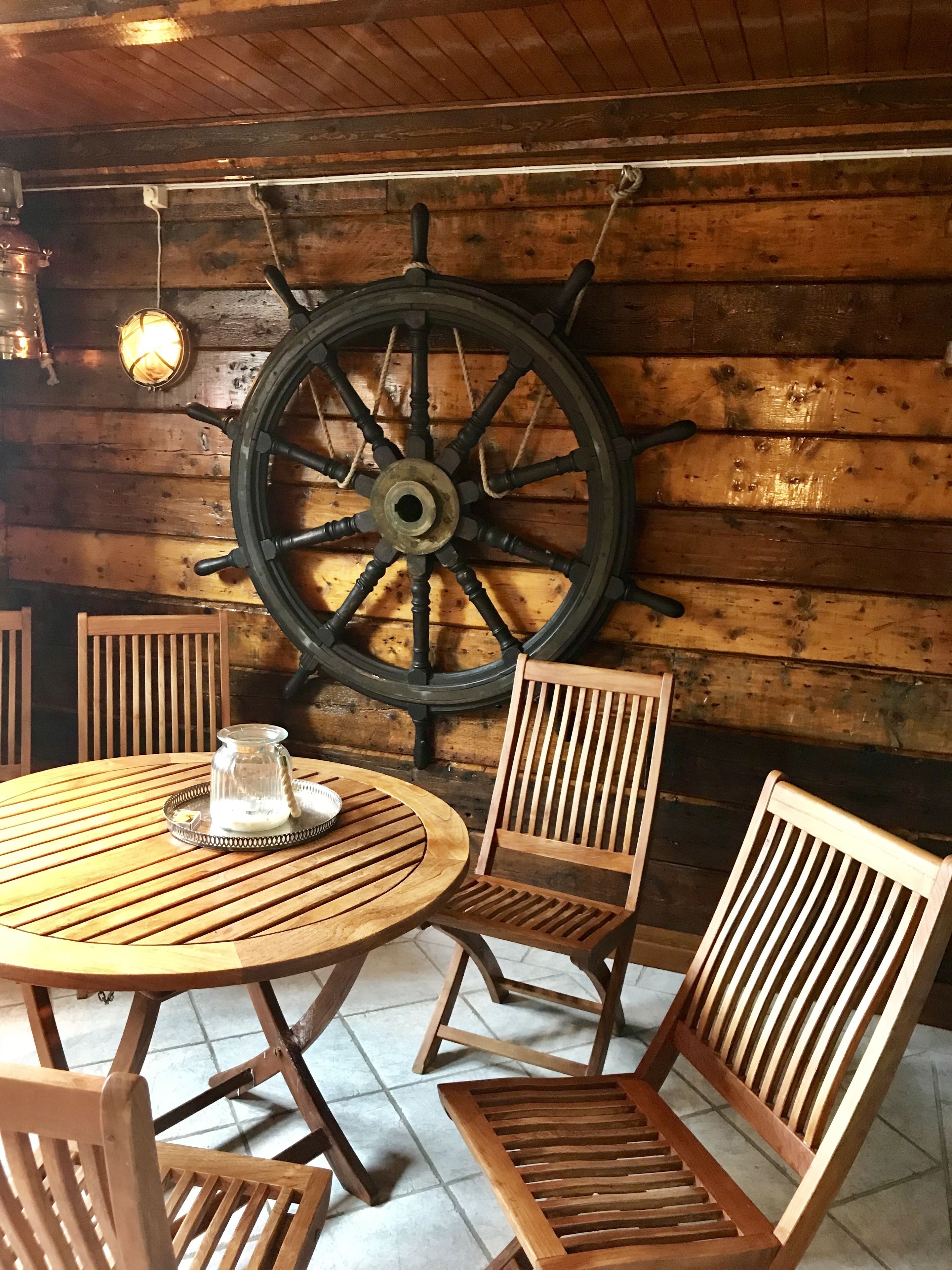 A wooden interior with a large ship's wheel mounted on the wall, a round wooden table with chairs, and a small glass pitcher on the table.