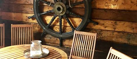 A wooden interior with a large ship's wheel mounted on the wall, a round wooden table with chairs, and a small glass pitcher on the table.
