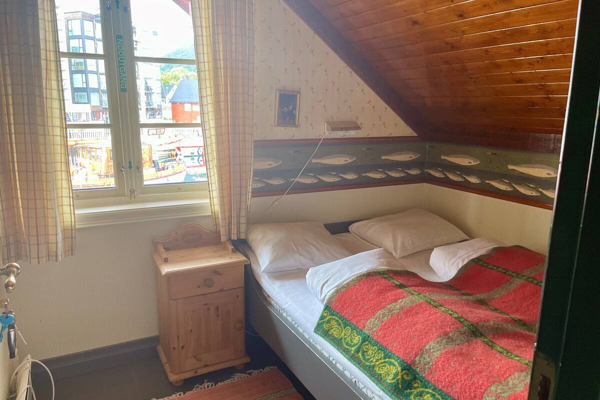 A bedroom with a bed, a nightstand, a window with curtains, and a view of buildings outside.