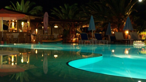A nighttime swimming pool with lounge chairs and umbrellas, surrounded by palm trees and a bar area. - Ninos on the Beach (Corfu)