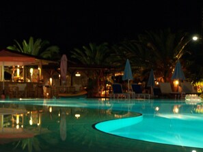 A nighttime swimming pool with lounge chairs and umbrellas, surrounded by palm trees and a bar area. - Ninos on the Beach (Corfu)