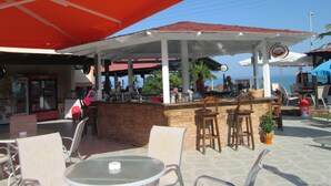 An outdoor bar area with tables and chairs, umbrellas, and a Coca-Cola refrigerator. - Ninos on the Beach (Corfu)