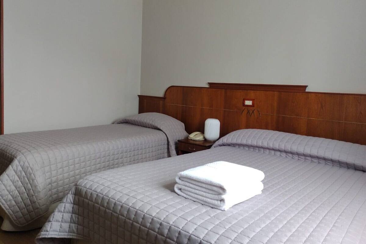 A hotel room with two beds, each with a quilted bedspread and pillows. There is a wooden headboard, a bedside table with a lamp, and a folded towel on one bed.