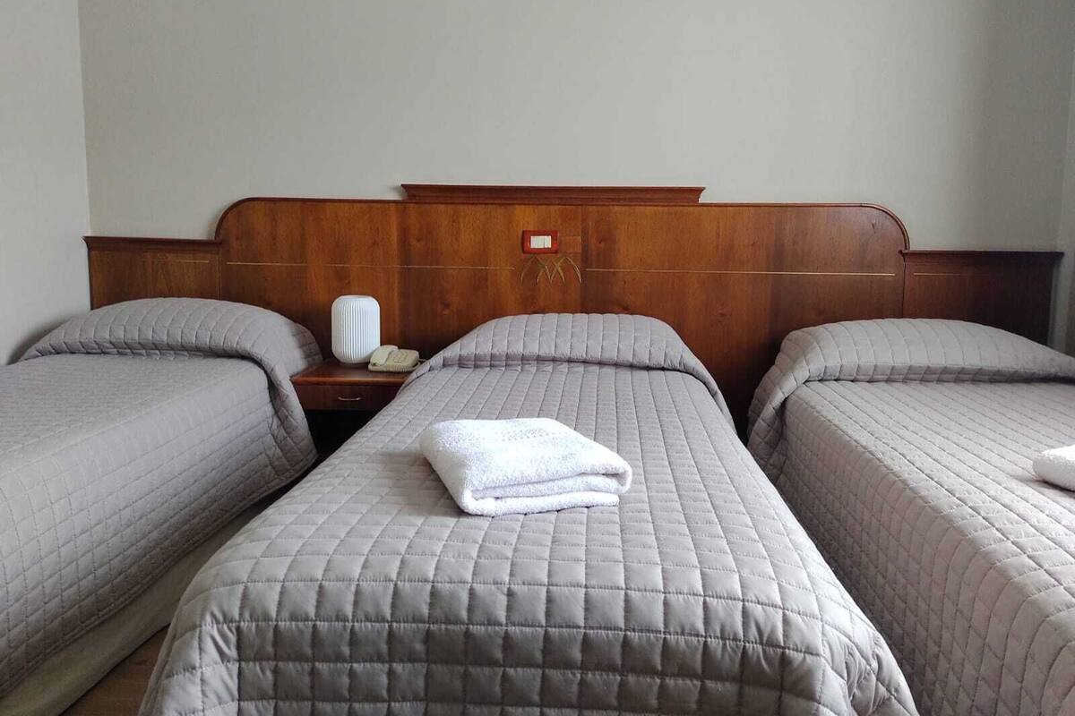 A hotel room with two single beds, each with a grey quilted duvet and pillows. There is a wooden headboard with a small red sign above the beds. A white bedside table with a lamp and a telephone is visible between the beds.