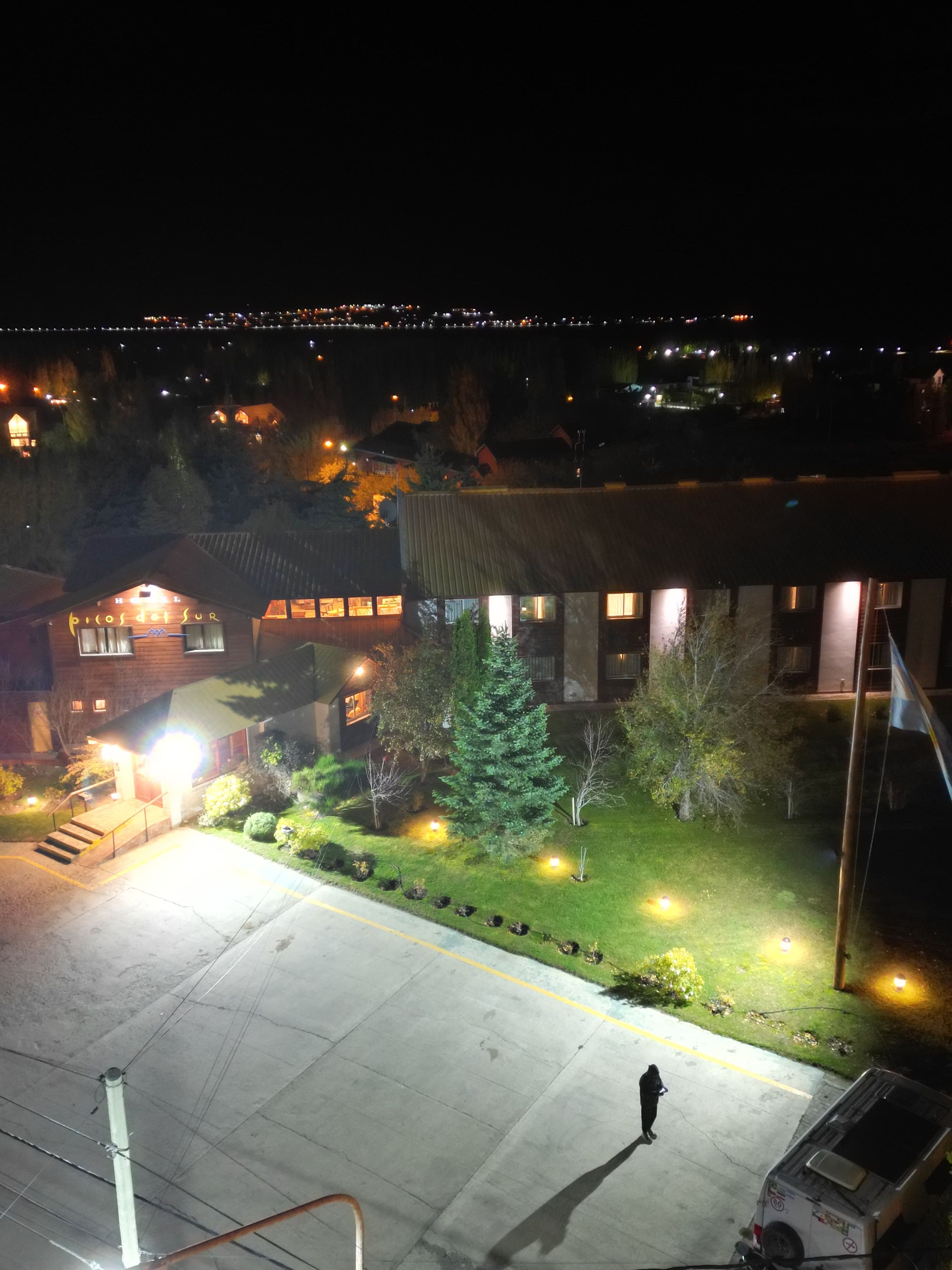 Um hotel com entrada bem iluminada, área de estacionamento e uma pessoa caminhando.