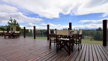 Una terraza de madera al aire libre con mesas y sillas, con vistas a un paisaje pintoresco con árboles y montañas.