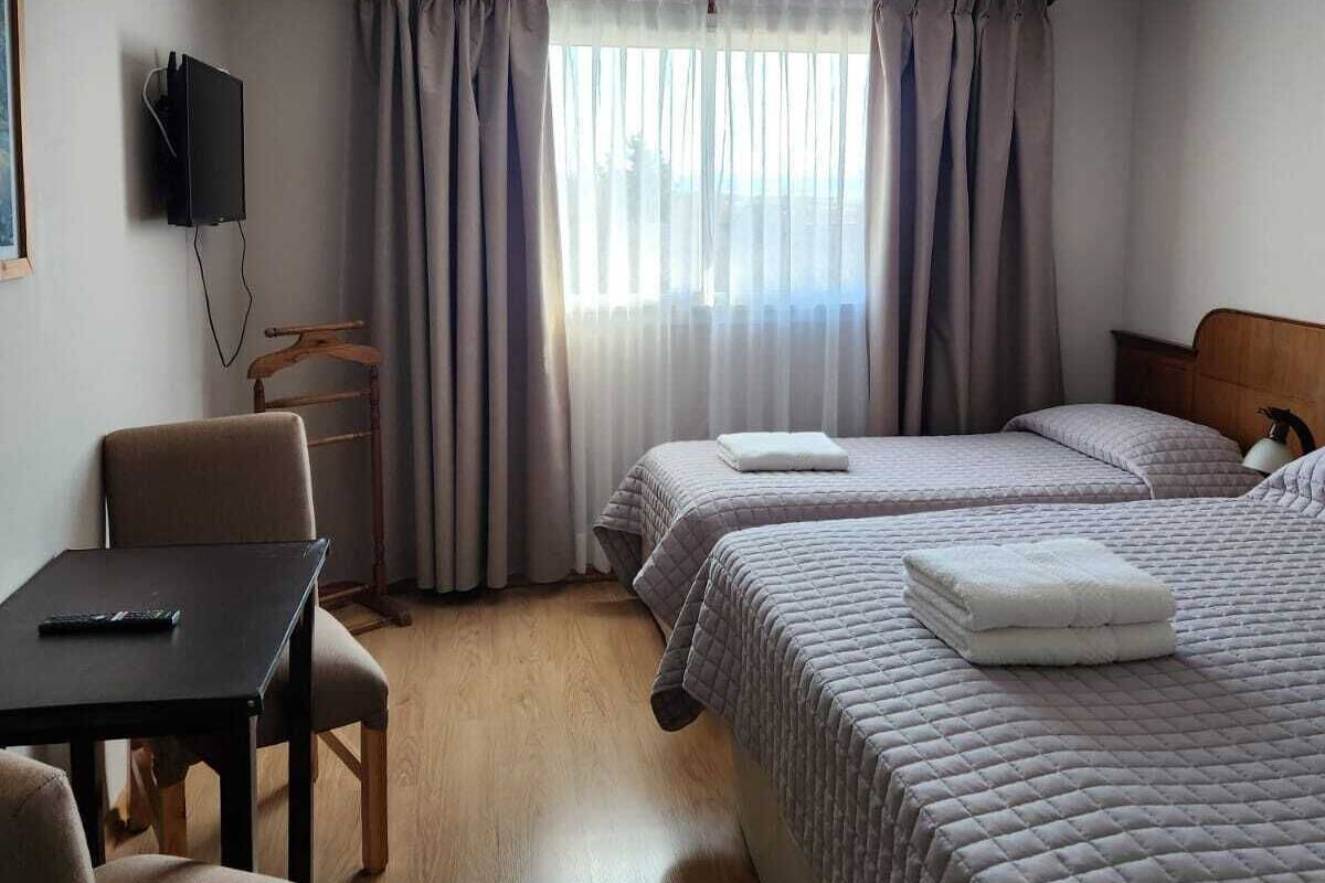 A hotel room with two beds, a desk, a chair, a TV, and a window with curtains.