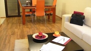 A modern living room with a wooden dining table, orange chairs, a black coffee table with papers and a bowl of fruit, and a white sofa with a black bag. - BIZ Apart Sofia (Sofia)