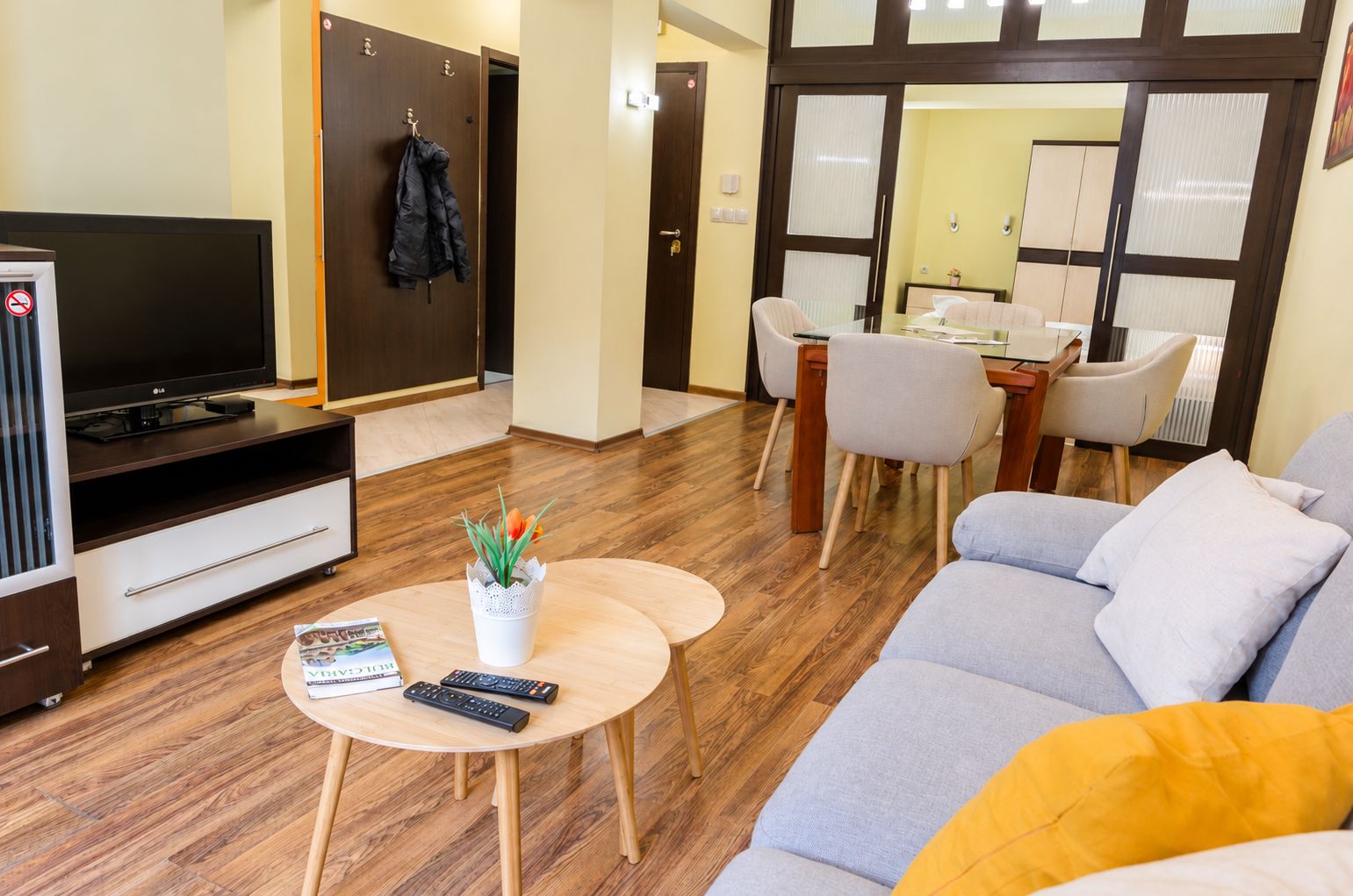 Una sala moderna con un televisor de pantalla plana, un sofá, una mesa de centro con mandos a distancia y un comedor con sillas y una mesa.
