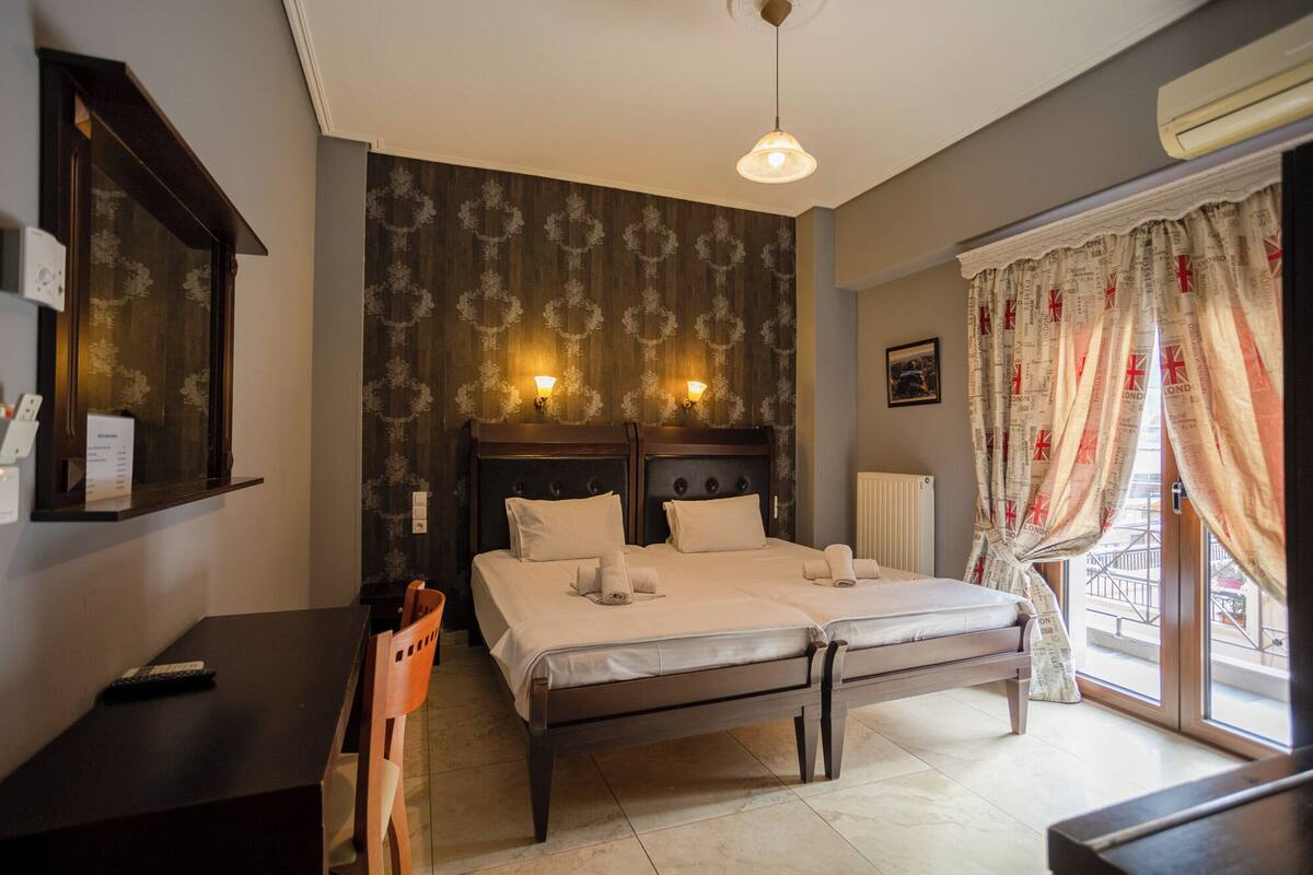 A double bed with a wooden headboard, a desk with a chair, a wall-mounted shelf, and a window with curtains.