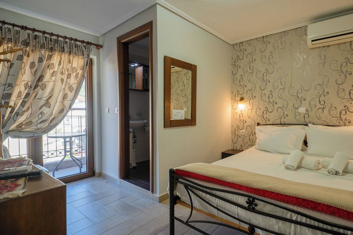 A hotel room with a bed, bedside tables, an air conditioning unit, a bathroom with a sink and mirror, and a view of the outside balcony.
