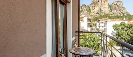 A balcony with a checkered table and chairs, a view of rocky mountains, and a white car parked nearby.