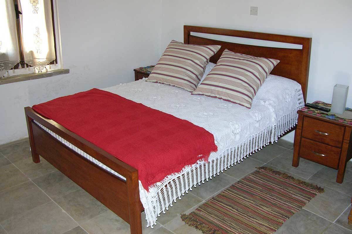 A bed with a red blanket, white lace bedspread, and striped pillows. There is a wooden nightstand with a lamp and a small drawer. A woven rug is on the floor.