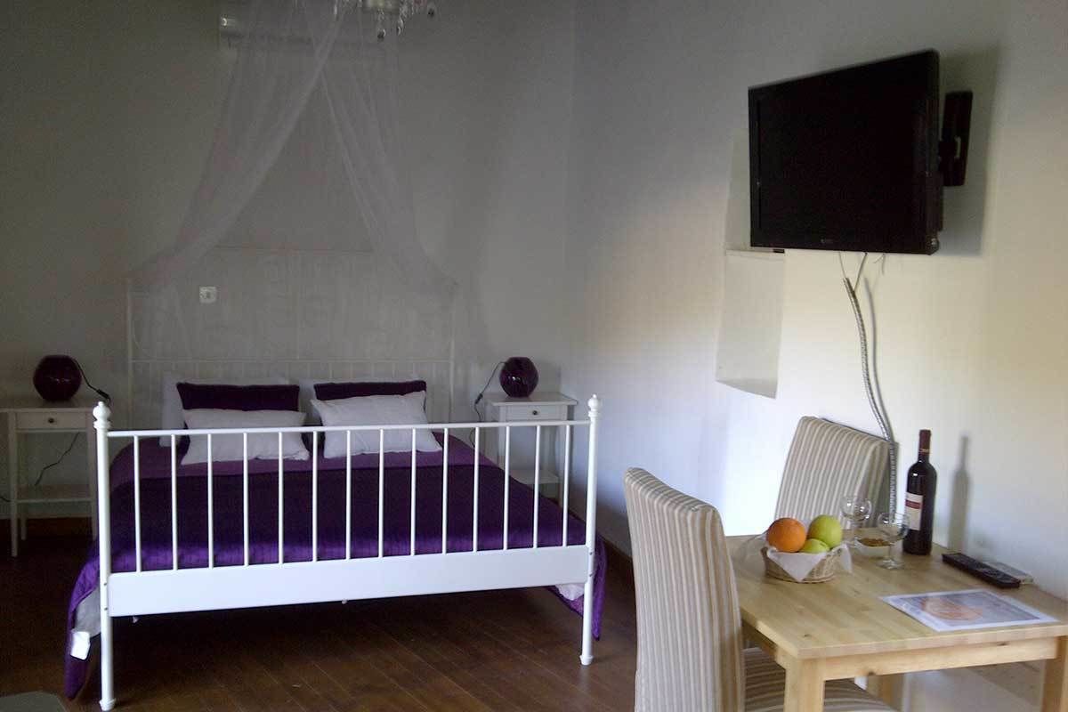 Una camera da letto con un letto bianco, una TV montata a parete, un comodino con una lampada e un tavolino con frutta e una bottiglia.